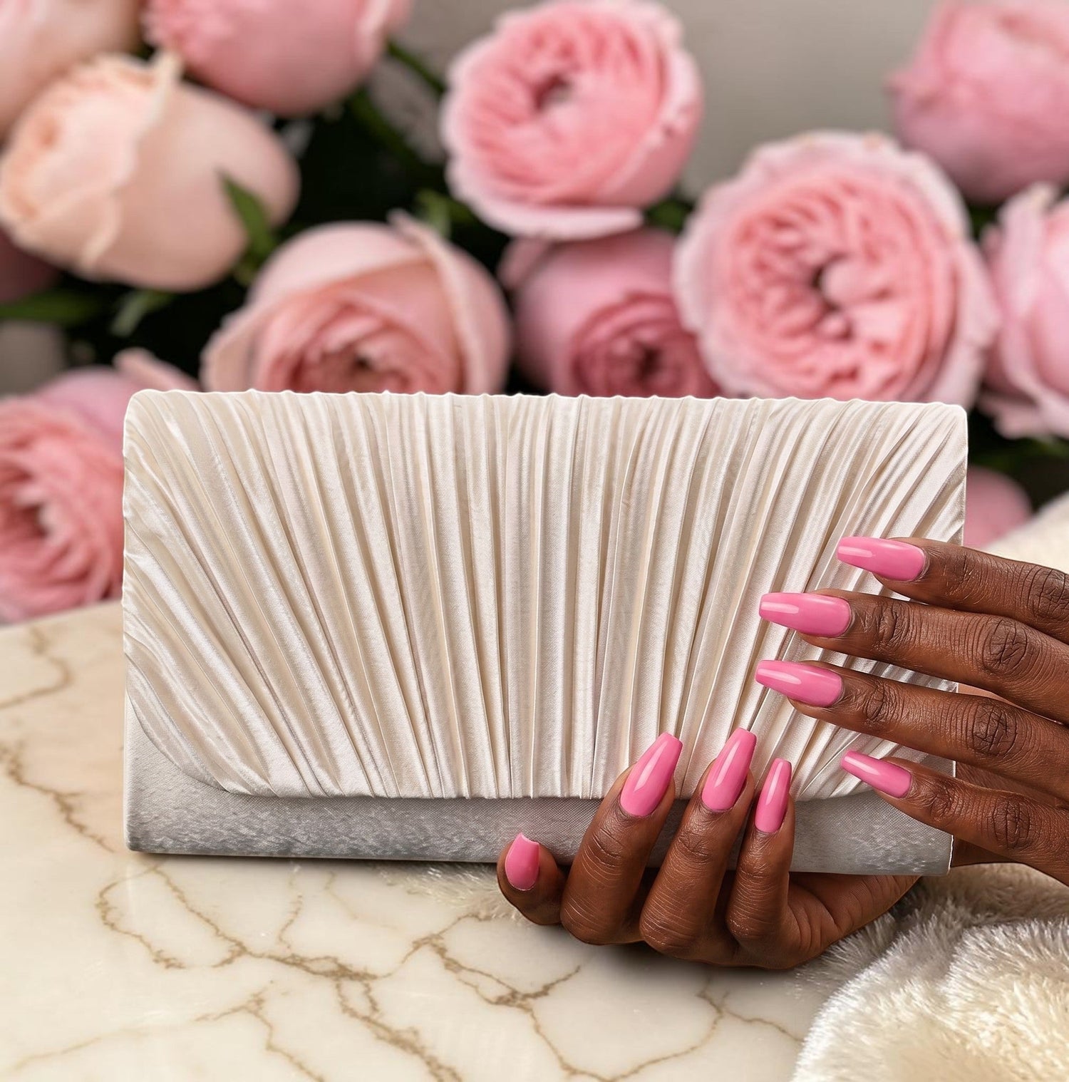 House of Lux Gifts cream pleated evening clutch held by hands with pink nails against a background of pink roses.