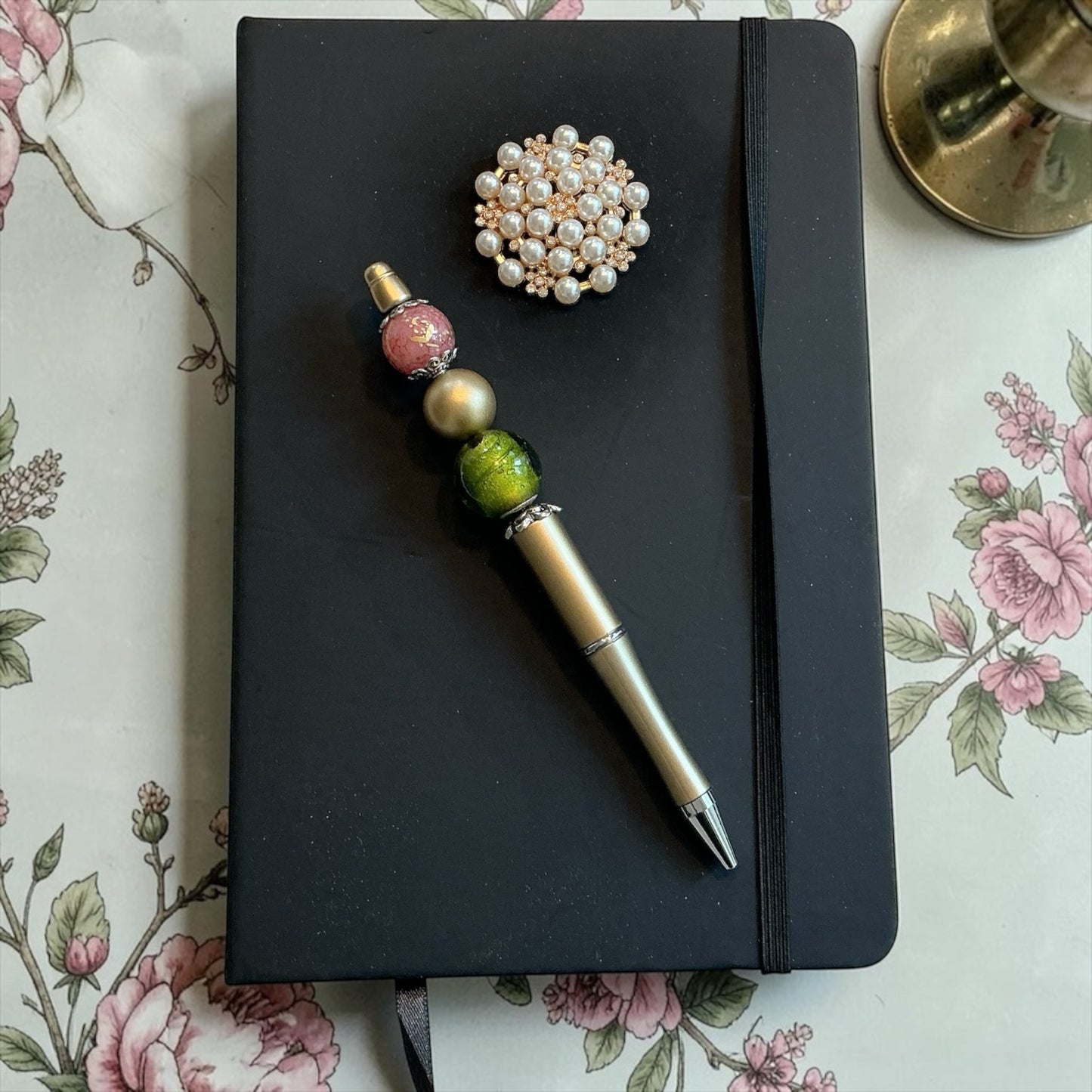 House of Lux Gifts beaded pen with pink, gold, and green beads resting on a black journal with a pearl brooch.