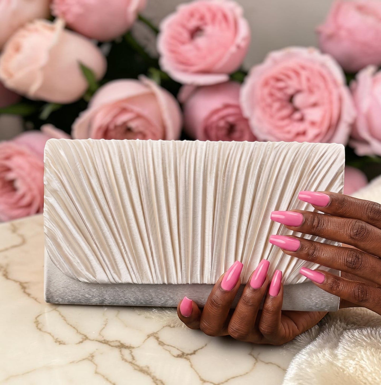 House of Lux Gifts cream pleated evening clutch held by hands with pink nails against a background of pink roses.