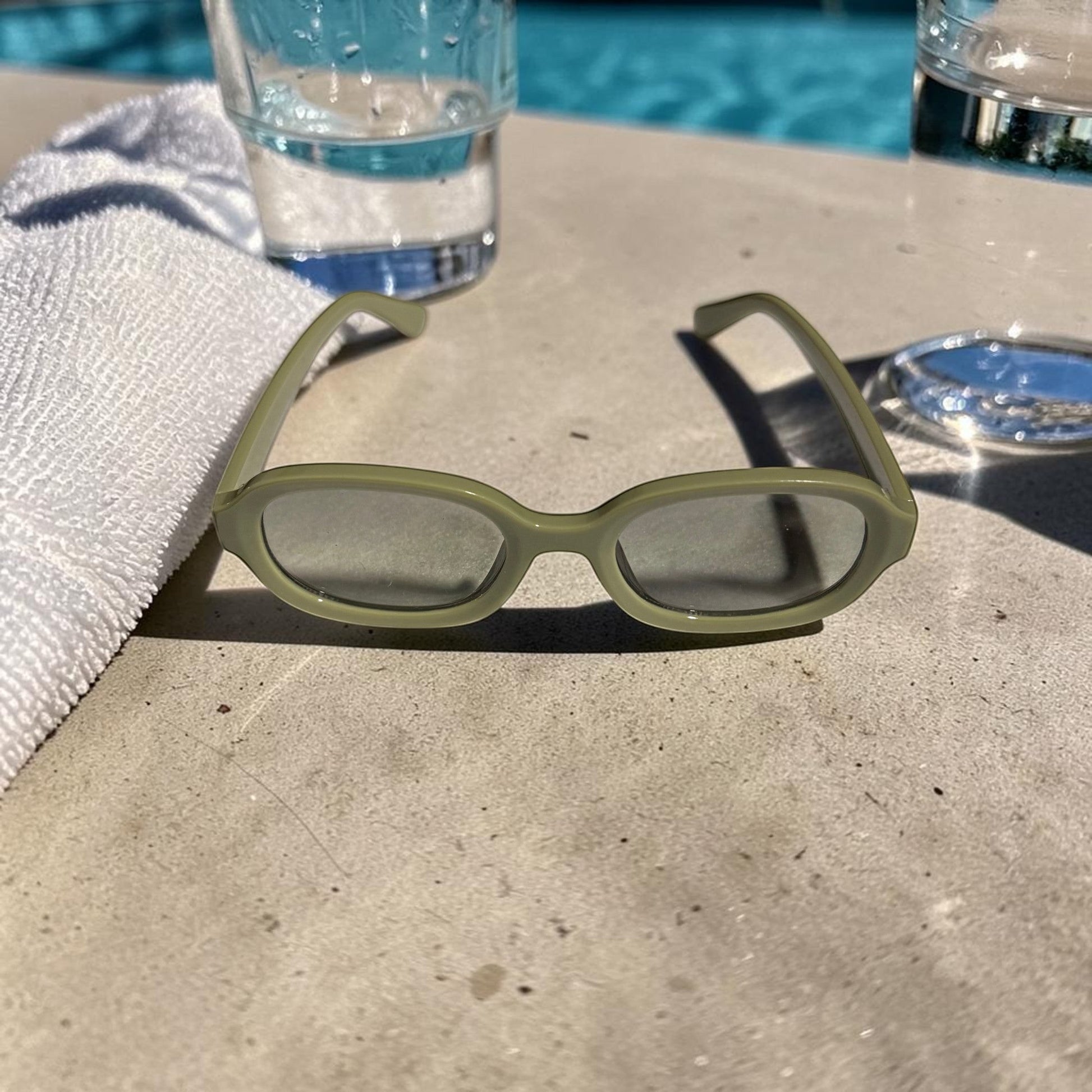 House of Lux Gifts olive green sunglasses with oval frames resting on a poolside ledge near a towel and glass.
