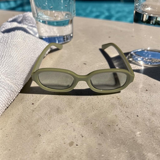 House of Lux Gifts olive green sunglasses with oval frames resting on a poolside ledge near a towel and glass.