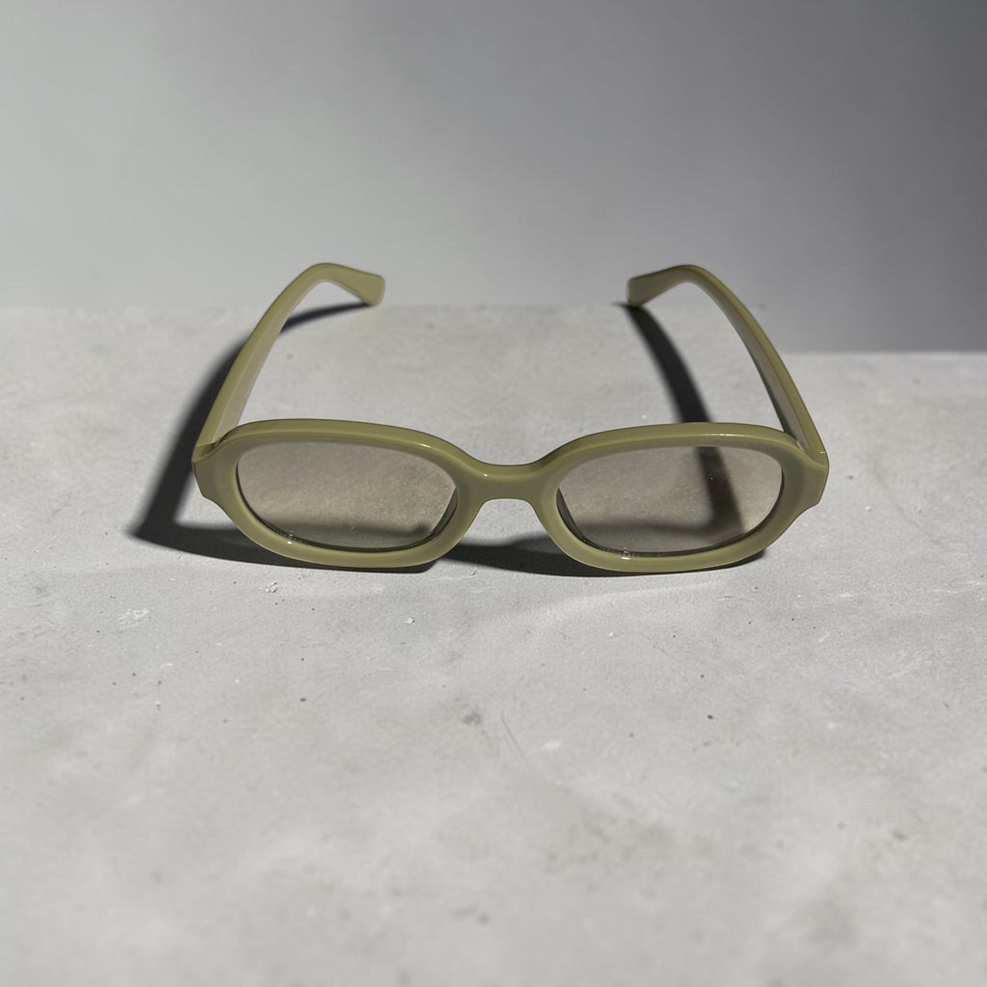 House of Lux Gifts olive green oval sunglasses with a minimalist frame design resting on a grey stone surface.